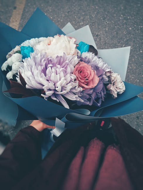 A hand-held bouquet of fresh flowers featuring large, soft lavender chrysanthemums, a pink rose, and smaller white and blue blooms, all arranged with green stems and leaves. The bouquet is wrapped in 
