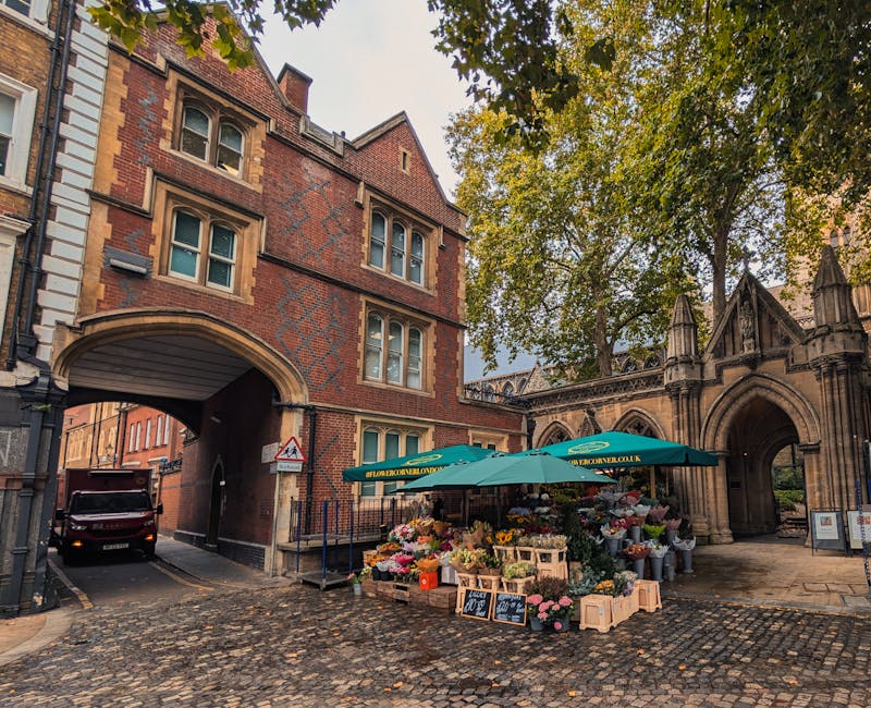 A bustling outdoor flower stall situated on a cobbled street under a large green umbrella, offering a variety of fresh flowers arranged in bouquets and crates. The flowers include pink roses, white li