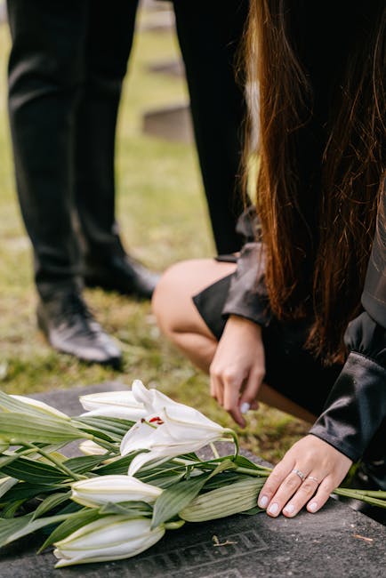 A person with long brown hair, dressed in dark clothing, is kneeling on a grave, placing a bouquet of white lilies with green leaves and long stems onto the grave marker. The bouquet is wrapped in lig
