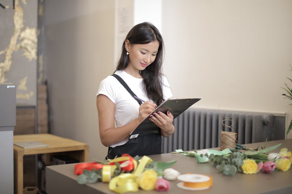 A young woman with long dark hair, wearing a white t-shirt and black apron, is standing at a table in a floral shop, writing notes on a clipboard. The table is adorned with various fresh flowers, incl