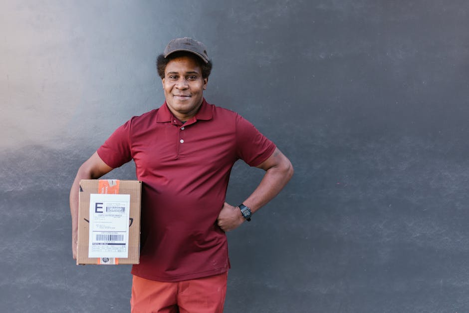 A smiling male courier with medium brown skin, wearing a maroon polo shirt, matching shorts, and a dark cap, stands in front of a dark grey wall. He holds a small rectangular parcel with an orange and