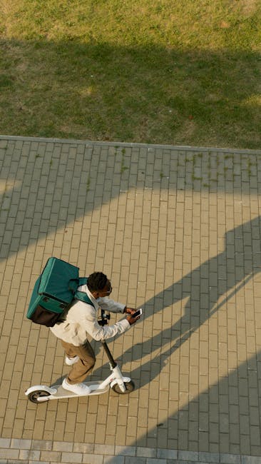 An individual riding a white electric scooter on a paved walkway, carrying a large, teal-colored delivery box attached to their back. The person is wearing a light-colored jacket and beige pants, with