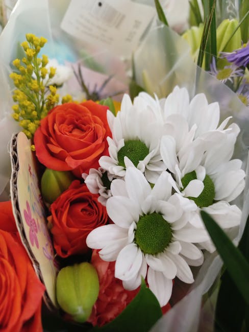 A close-up of a floral arrangement featuring white daisies with bright green centers, complemented by vibrant orange roses with layered petals, and small yellow filler flowers. The bouquet is wrapped 